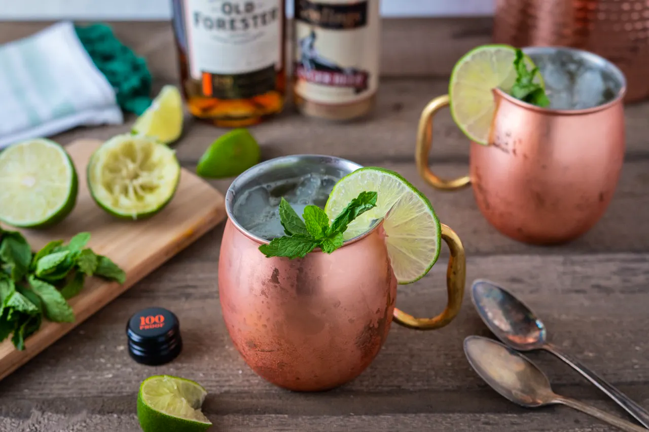 Two copper Moscow mule mugs garnished with lime and mint beside a cutting board of limes and bourbon bottles