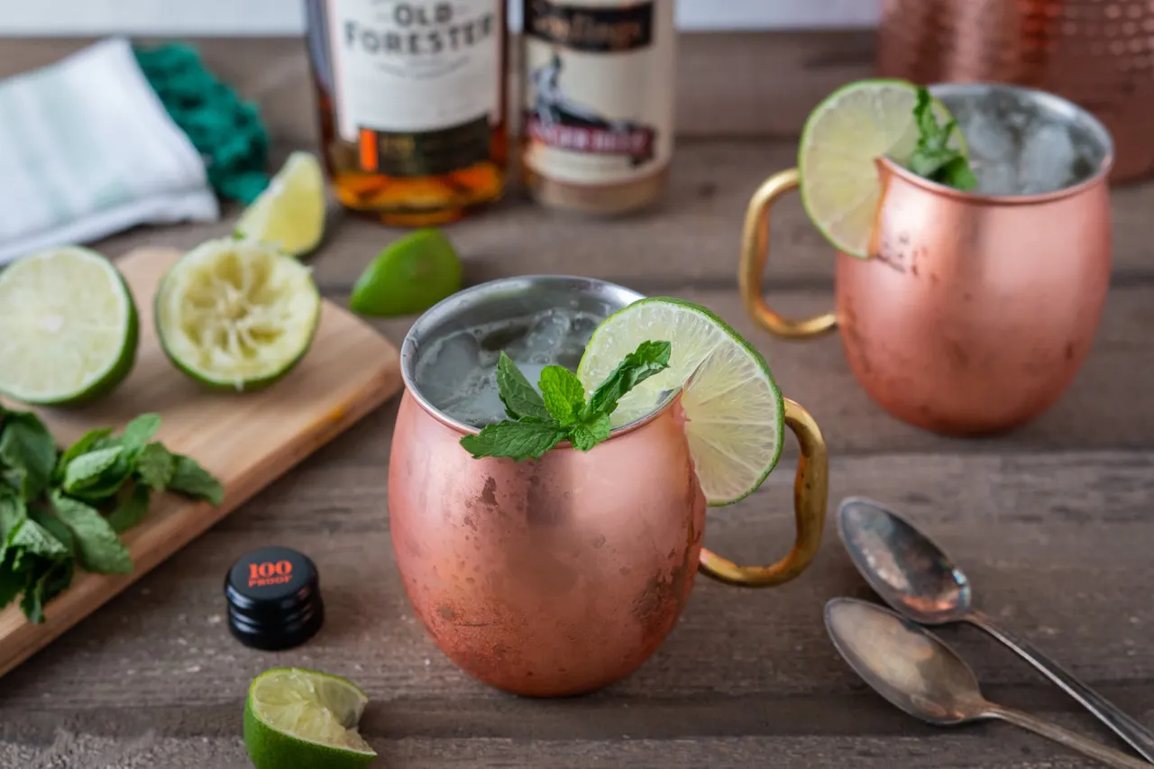 Two copper Moscow mule mugs garnished with lime and mint beside a cutting board of limes and bourbon bottles