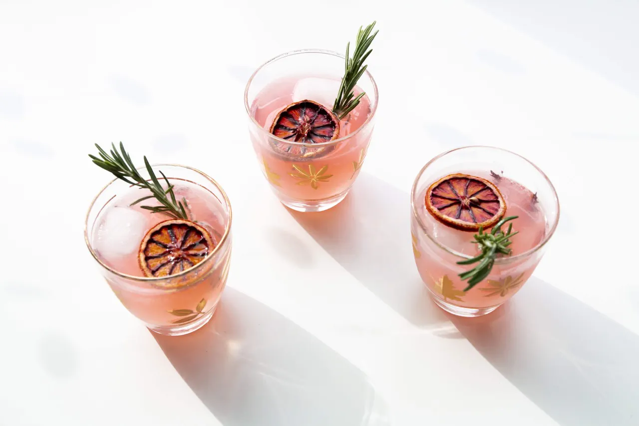 Three pink cocktails in gold-patterned glasses garnished with rosemary and dried blood orange slices