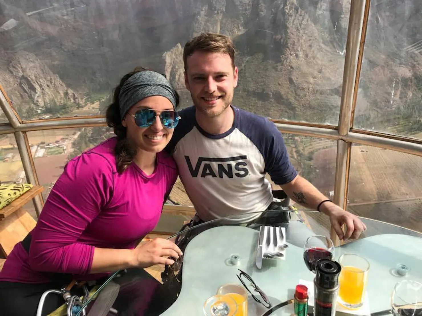 Two smiling people dining at a table inside a glass pod suspended on a cliff above a mountain valley