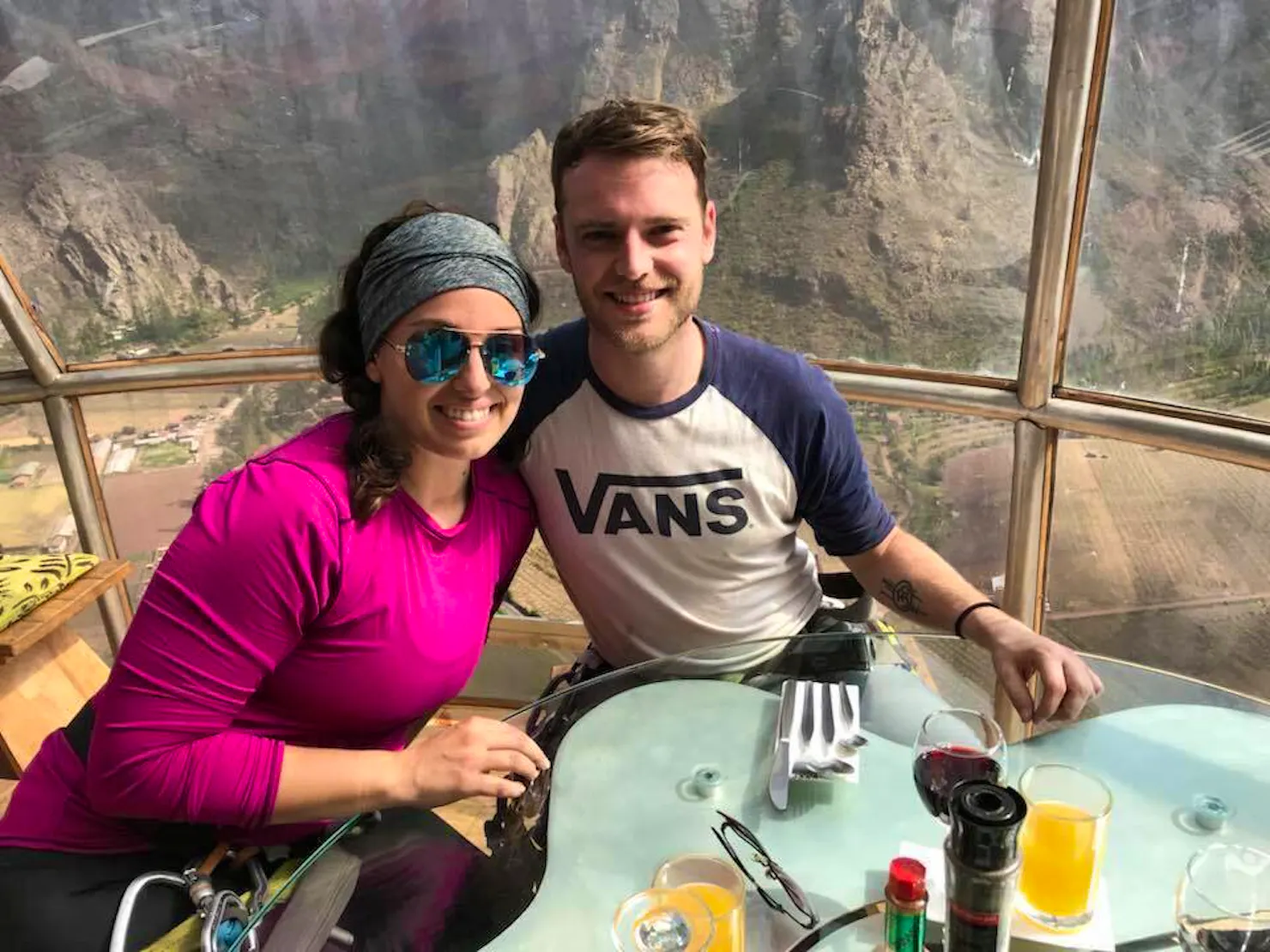 Two smiling people dining at a table inside a glass pod suspended on a cliff above a mountain valley