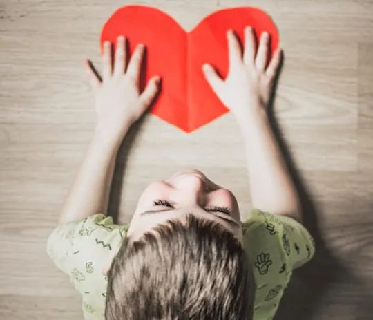 Child seen from above holding a red paper heart on a wooden floor, for the Necco project