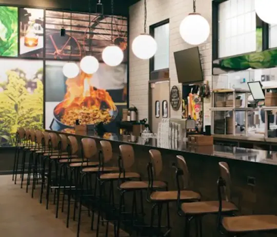 Kitchen 1883 restaurant interior with a long bar, wooden stools, globe pendant lights and a flaming-skillet mural