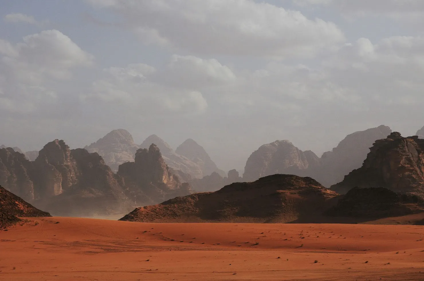 Red sand desert with rugged rock mountains under a hazy, cloudy sky
