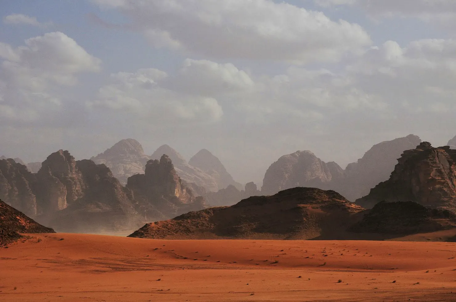 Red sand desert with rugged rock mountains under a hazy, cloudy sky