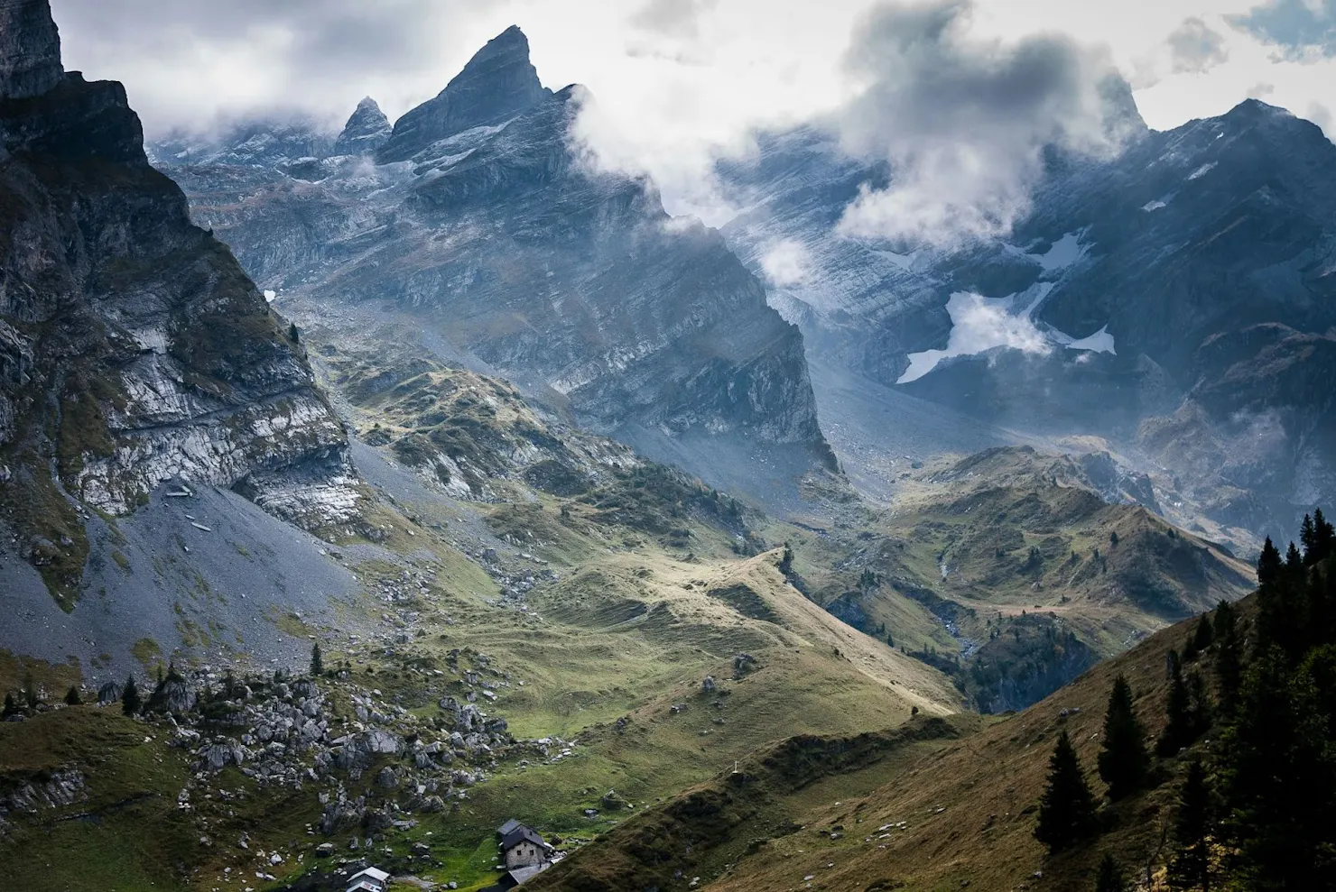 Misty alpine valley with jagged peaks, green slopes and a small cabin in the foreground