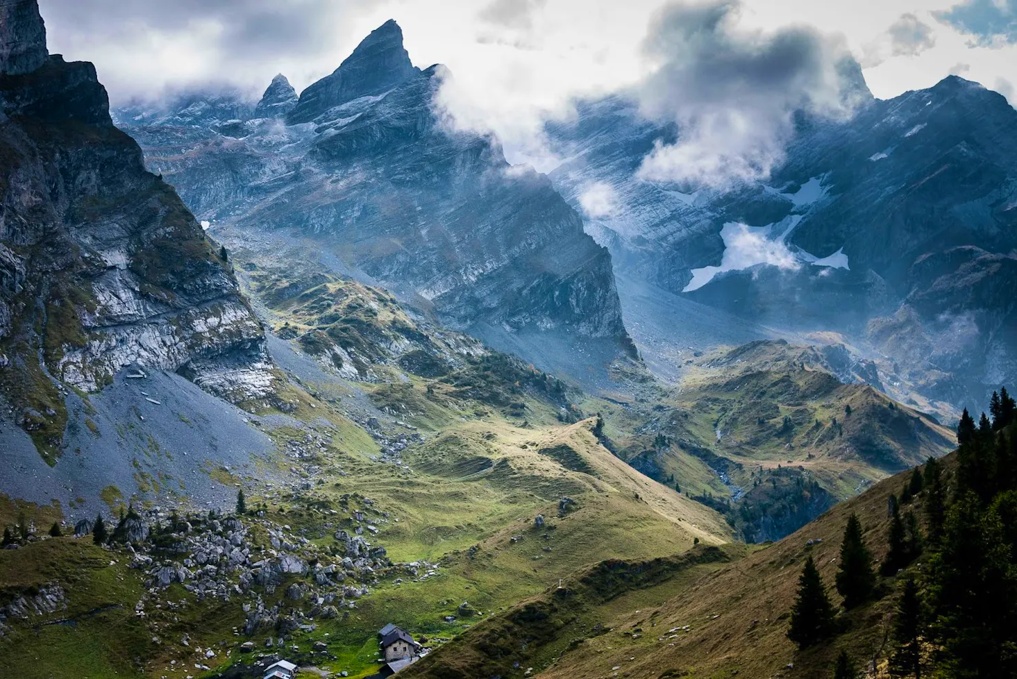 Misty alpine valley with jagged peaks, green slopes and a small cabin in the foreground