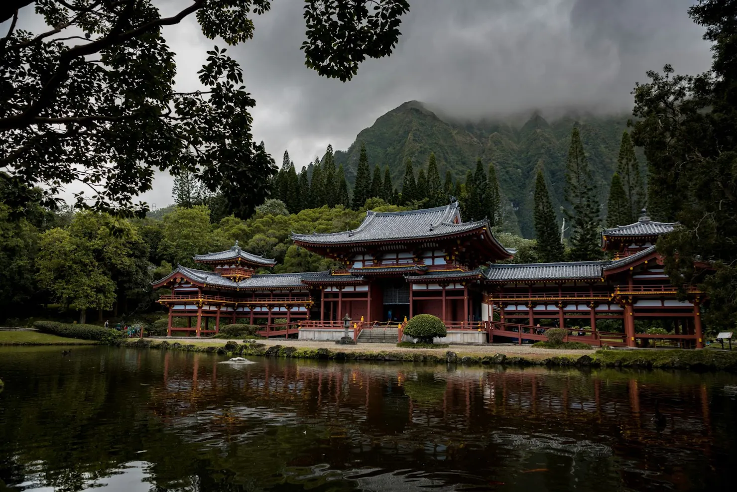 Red and white temple reflected in a pond, with forested mountains and low clouds behind