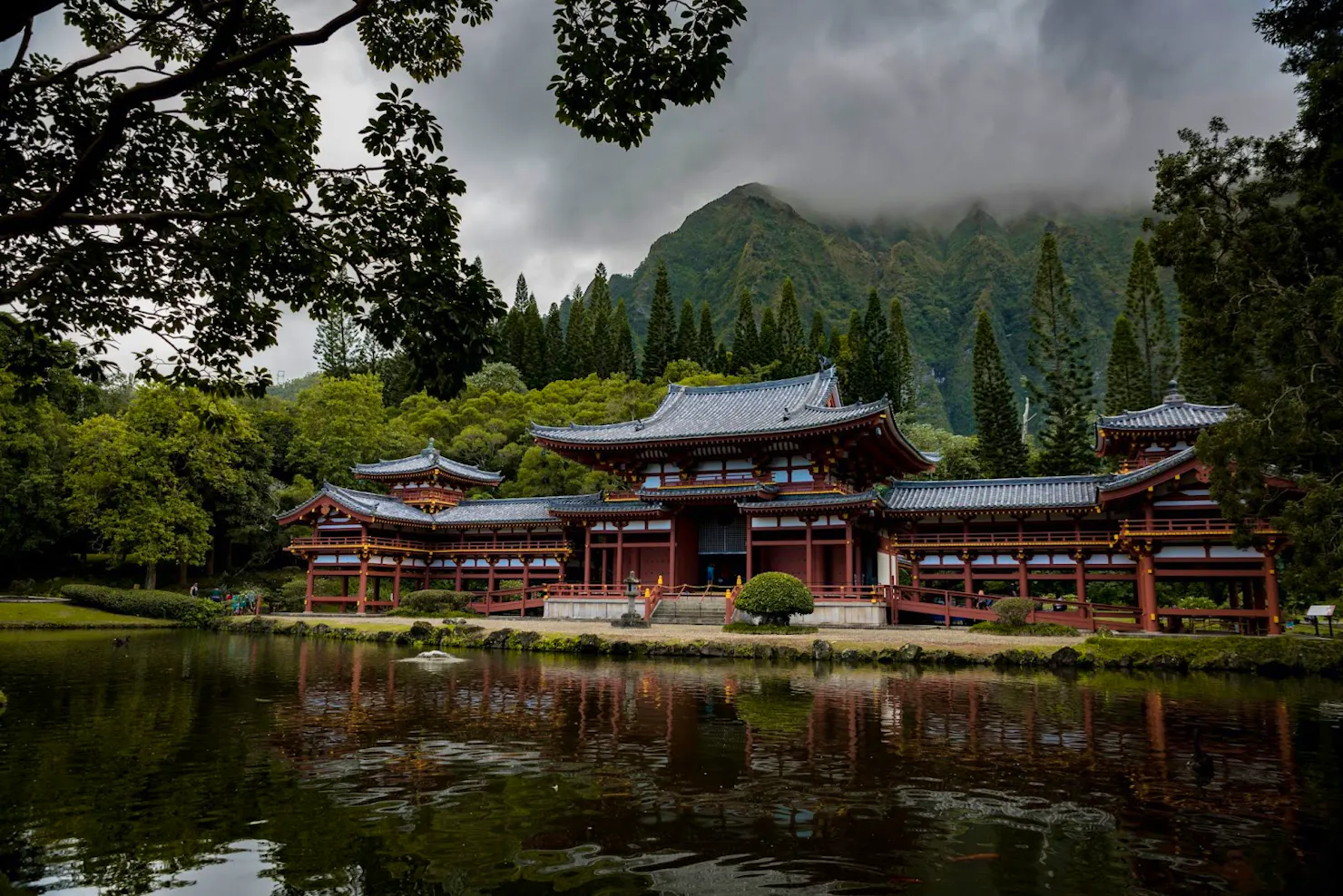 Red and white temple reflected in a pond, with forested mountains and low clouds behind