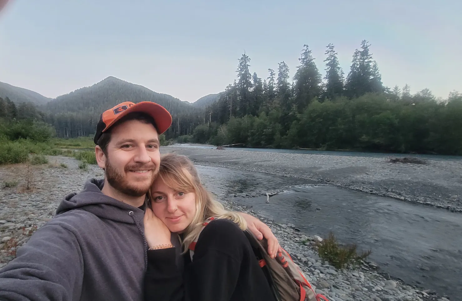 Ben Iburg and a companion taking a selfie beside a river with forested mountains behind them