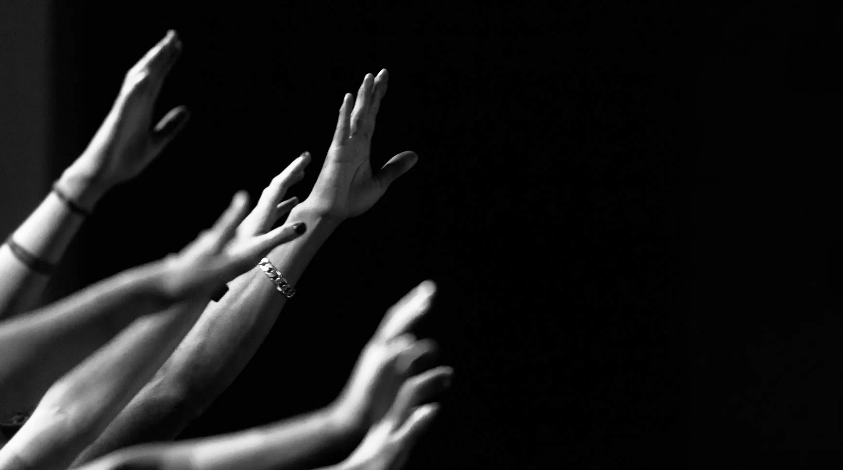 Black-and-white photo of raised hands reaching upward in worship against a dark background
