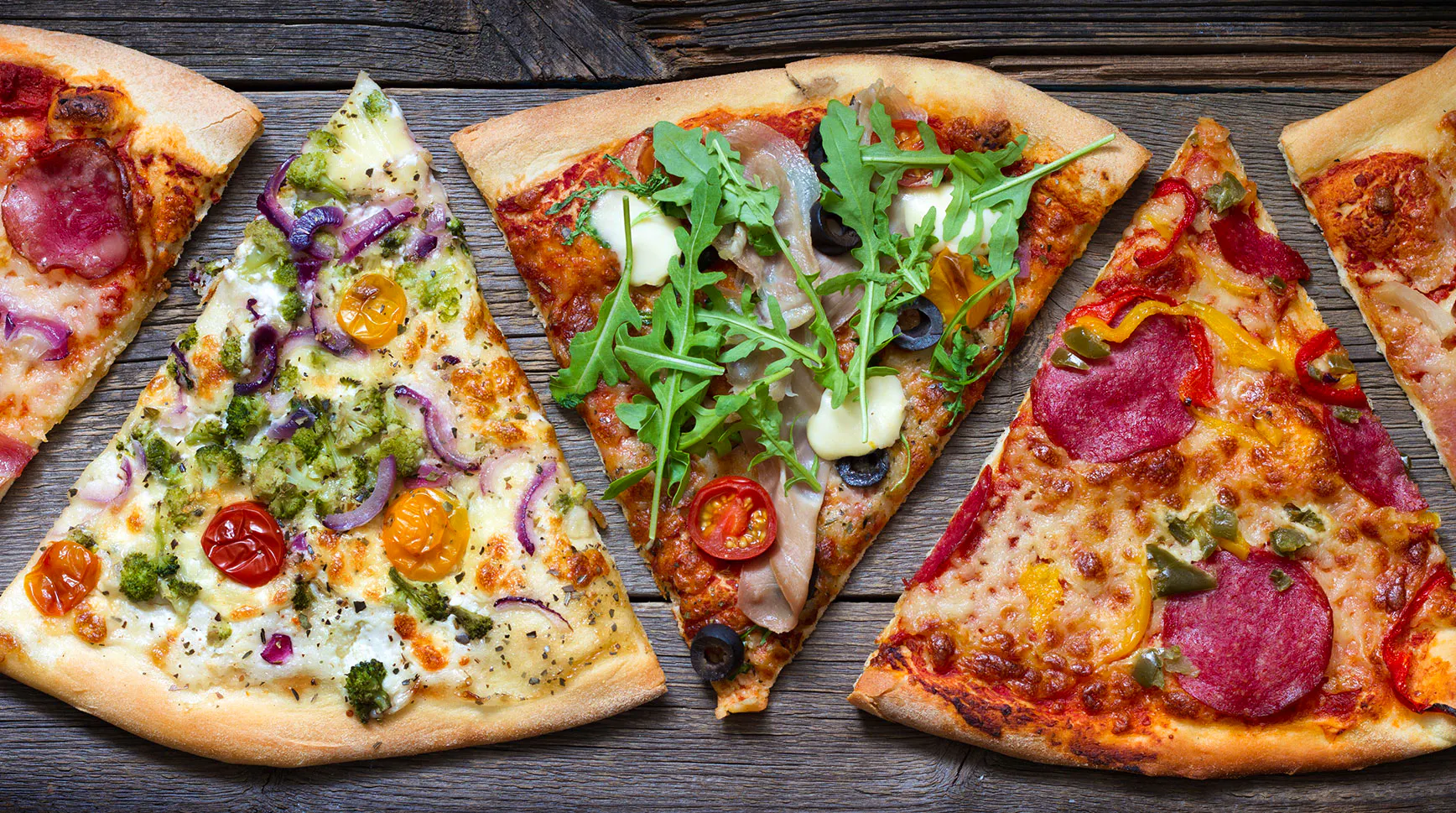 Pizza slices with different toppings, from prosciutto and arugula to salami and peppers, laid out on a wooden table
