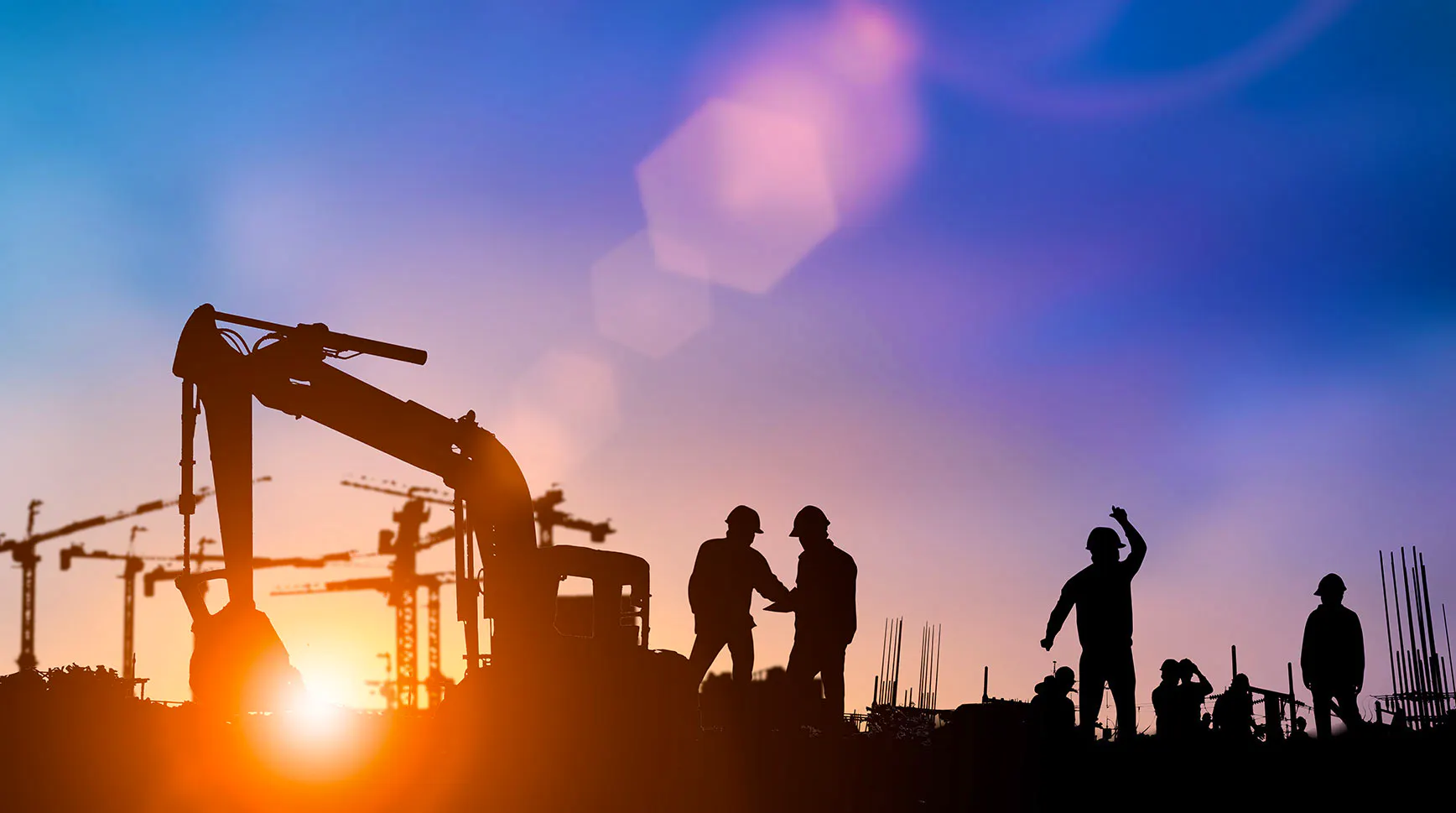 Silhouettes of construction workers and an excavator against a sunset sky with cranes in the background
